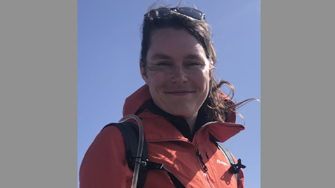 Lächelnde Frau in roter Outdoorjacke mit Rucksack vor blauem Himmel.