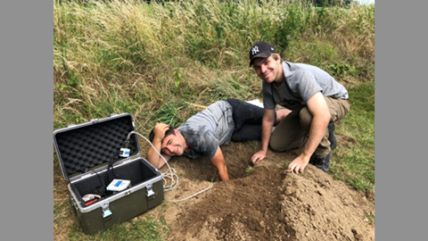 Zwei Männer bei archäologischen Grabungen mit technischem Messgerät im Feld.