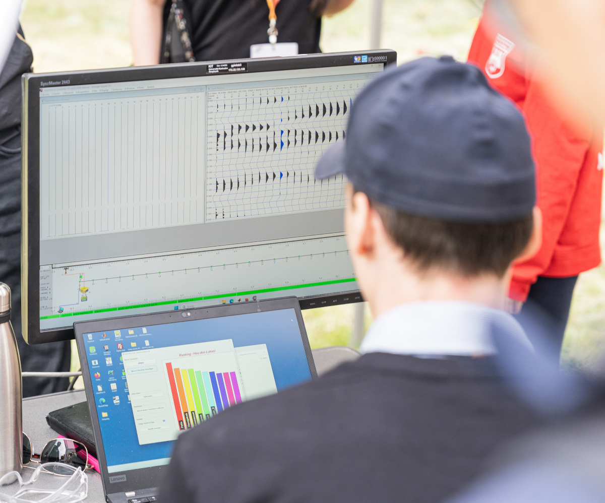 Mann mit Kappe analysiert wissenschaftliche Daten auf einem Monitor und einem Laptop im Freien.
